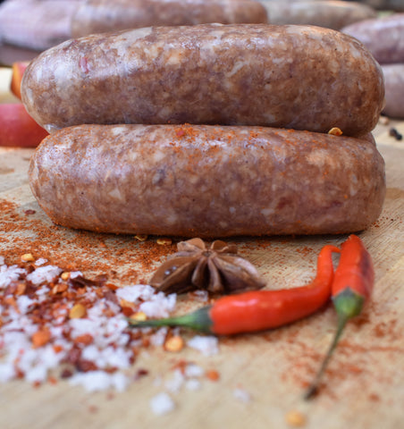 Three un-cooked Firecracker sausages placed on an old farmhouse table, with red chillie peppers and other ingredients
