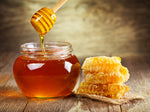 A round jar containing golden clear honey. A honey drizler is lifting some honey from the jar, some of which is flowing back into the jar. The jar is sat on a wooden table, next to honeycomb.